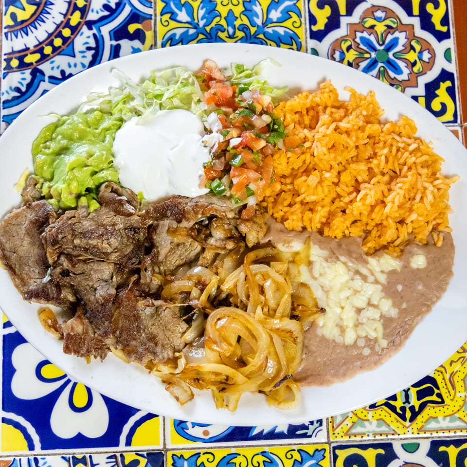 A white plate of carne asada with rice, beans, guacamole, sour cream, and pico de gallo.