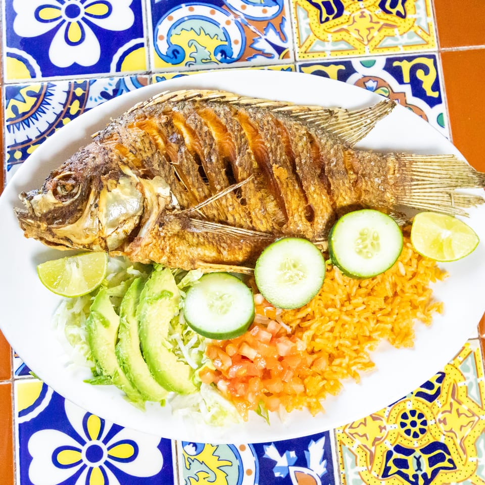 A whole fried fish with rice, avocado, lettuce, cucumber, tomato, and lime on a white plate.