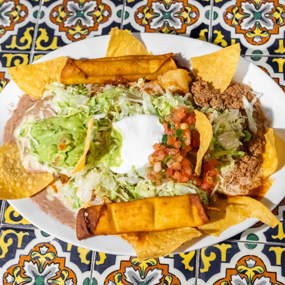 A white plate of Mexican food with taquitos, nachos, guacamole, sour cream, and salsa on a colorful tiled table.