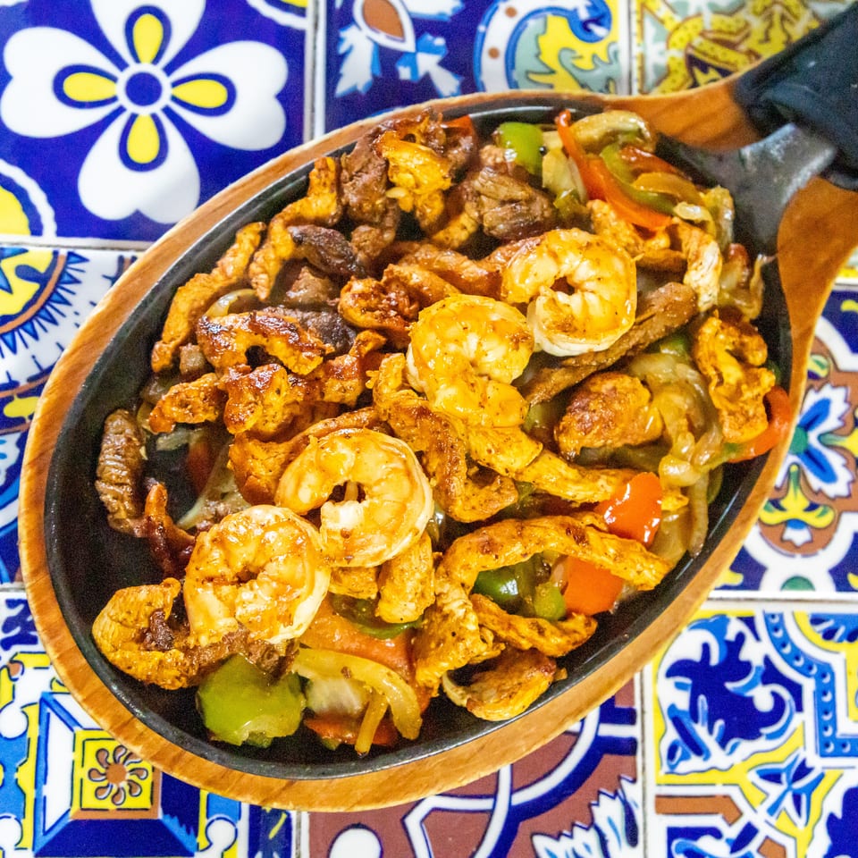 Sizzling fajitas with chicken, steak, shrimp, and colorful vegetables on a decorative tiled table.