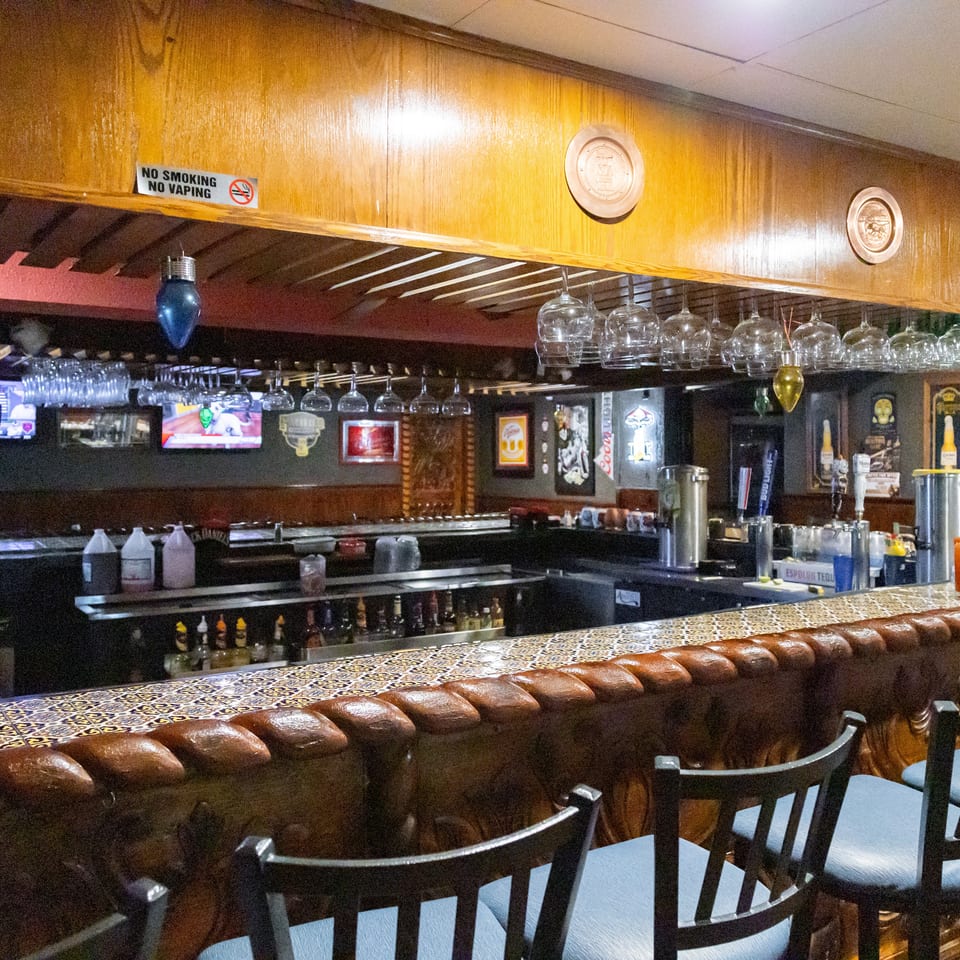 A long, dark wood bar with blue-seated stools, hanging glasses, and a "No Smoking" sign.