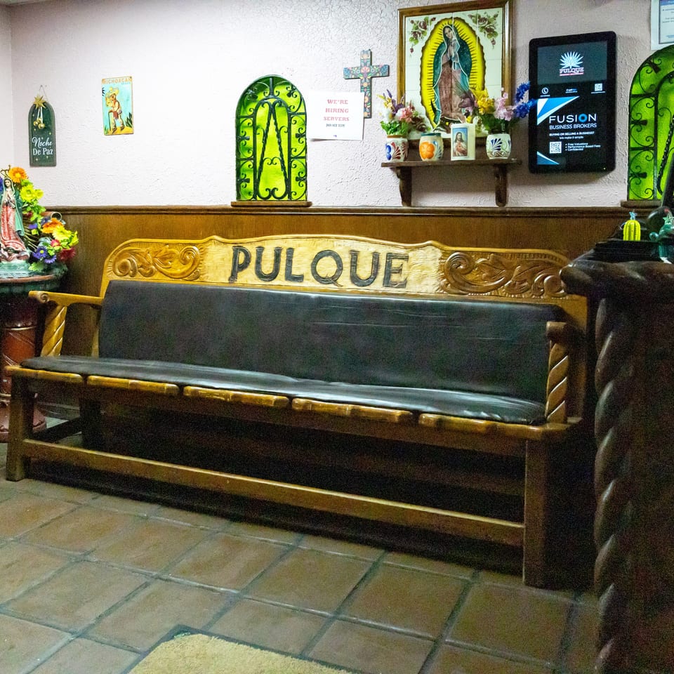 A wooden bench with "PULQUE" carved into its back, surrounded by religious decor and restaurant signs.