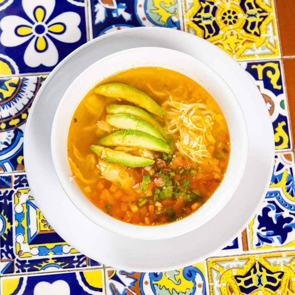Chicken tortilla soup with avocado slices and cilantro, served on a colorful tiled table.